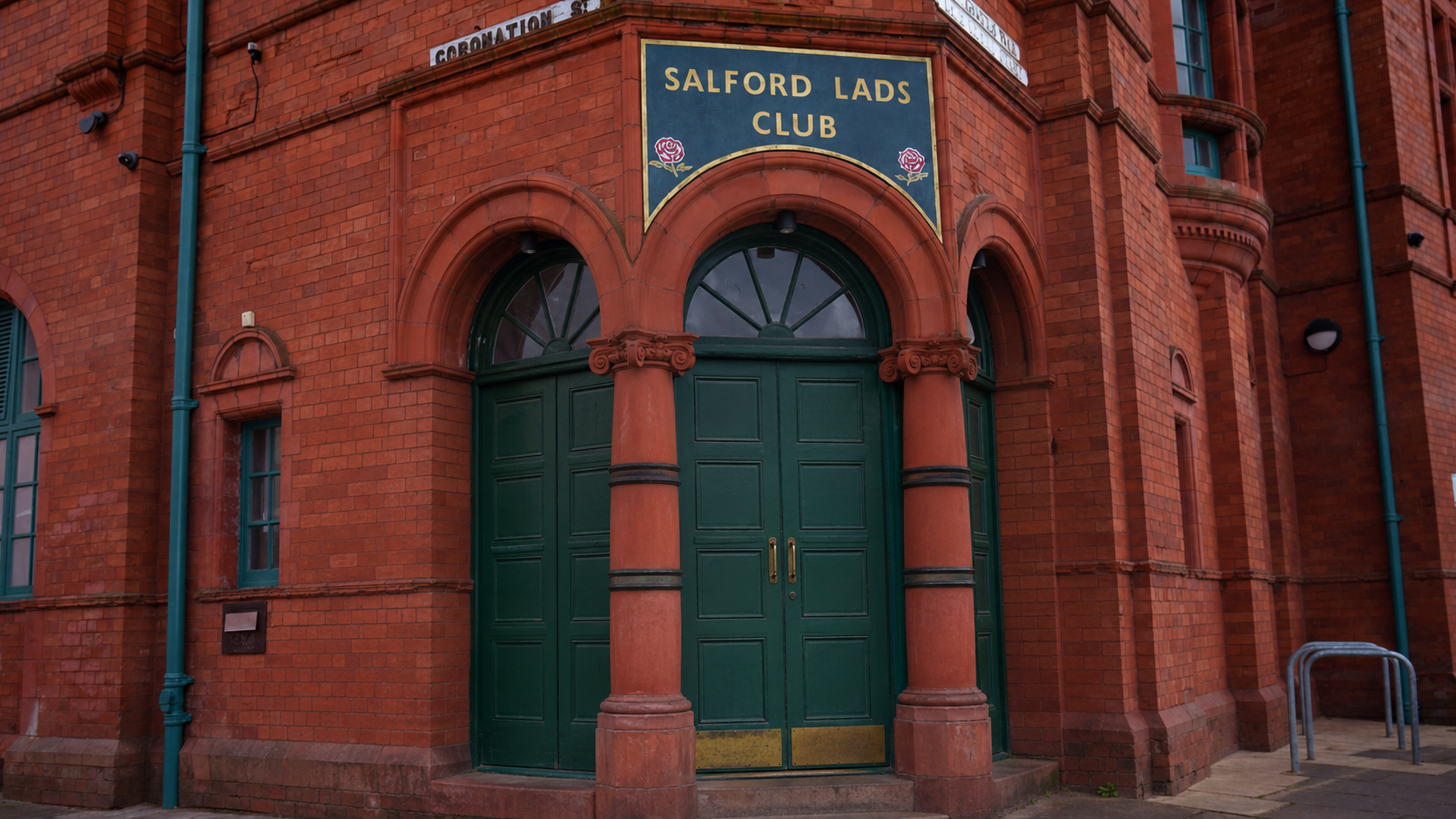 Red-brick building entrance with arched green double doors and columns, topped by a sign reading “Salford Lads Club.”
