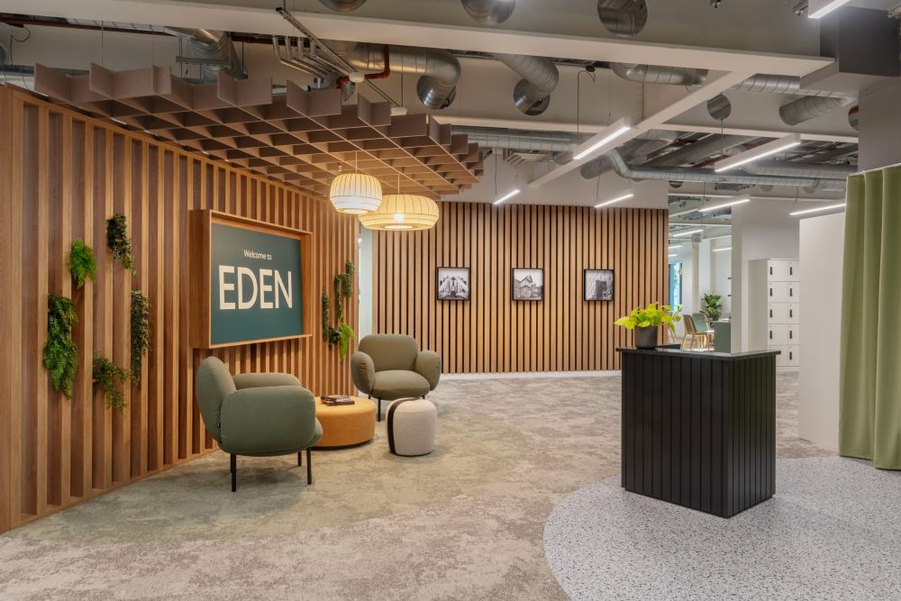 Welcoming office reception area with wood‑slat walls, green lounge chairs, hanging lights, plants, and a sign reading ‘Welcome to Eden,’ set beneath an exposed ceiling