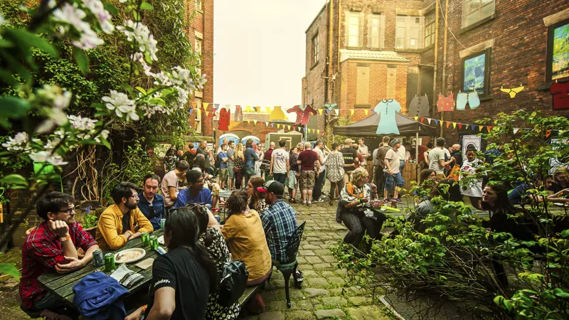 Gobbled courtyard area with people sat on wooden benches, standing around, colourful bunting and a fake washing line of clothes are hung up.