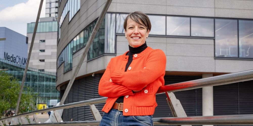 Alice Webb standing outdoors on a pedestrian bridge with arms folded, wearing an orange cardigan over a black high-neck top and blue jeans. Modern office buildings with large windows form the background, including a building displaying the text “MediaCityUK.” Metal railings and bridge supports frame the foreground.